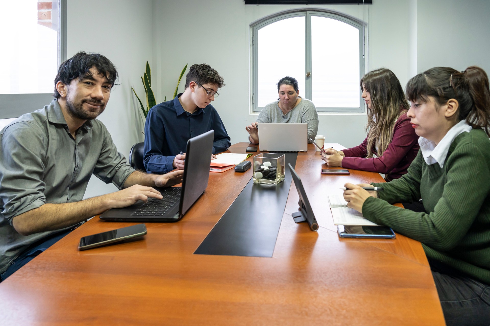 Grupo de empleados sentados alrededor de la mesa de reuniones de la oficina trabajando en sus cuadernos, uno de ellos mirando a la cámara