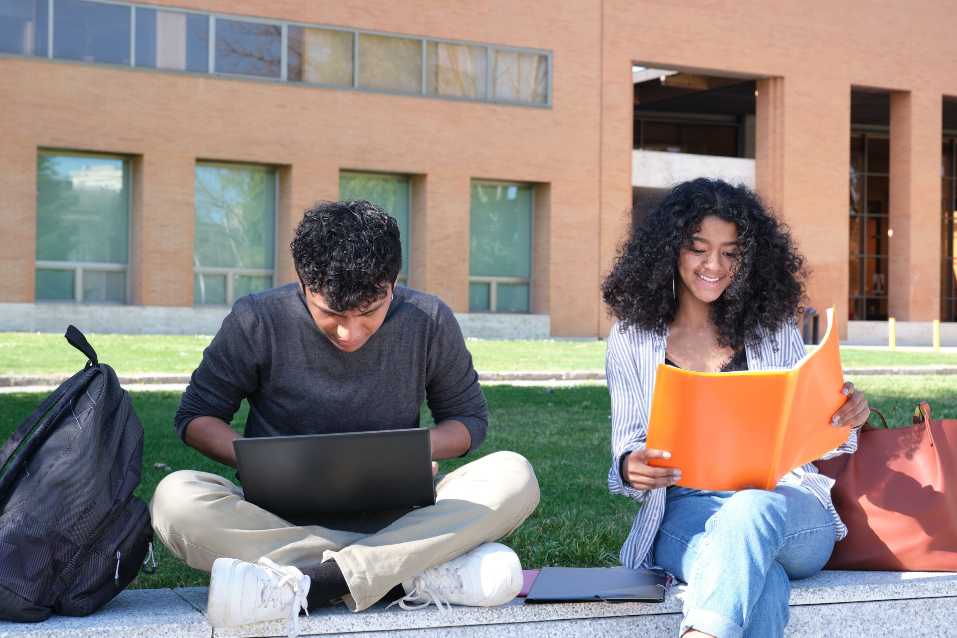 Dos estudiantes de latín estudiando desde sus notas de clase sentados en una pared al aire libre. Vida universitaria en el campus.